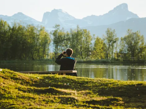 En person sitter på en benk og ser i en kikkert på fjellene i bakgrunnen.