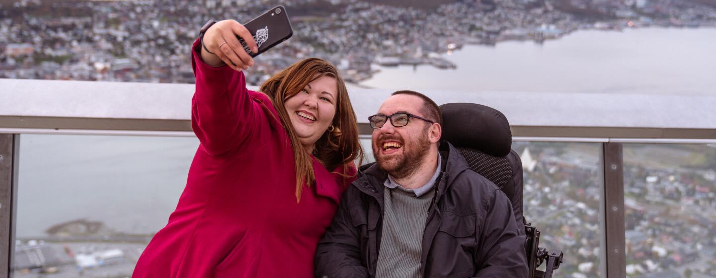 En mann i rullestol og en dame tar selfie oppe på Fjellheisen i Tromsø